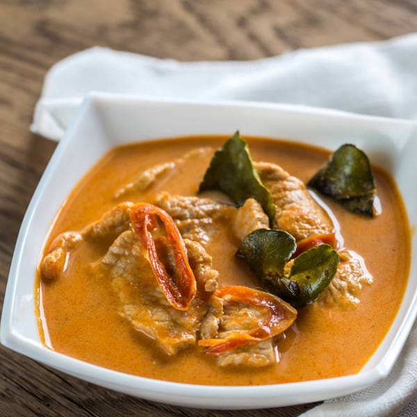 Burmese curry with meat, red peppers, and green leaves in a white bowl on a wooden table.